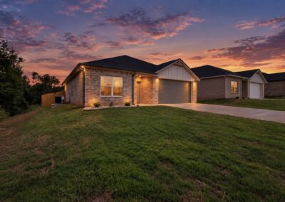 A single-story brick house with a two-car garage, illuminated at dusk. The sky is colorful with purple and orange hues. The home is surrounded by a well-kept lawn and a concrete driveway.