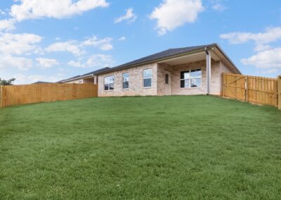 A modern brick house with large windows sits behind a wooden fence, surrounded by a spacious, well-maintained green lawn under a partly cloudy blue sky.