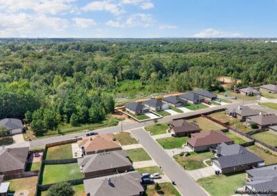 Aerial view of a suburban neighborhood with houses, fenced yards, streets, and driveways, bordered by a dense forested area under a partly cloudy sky.