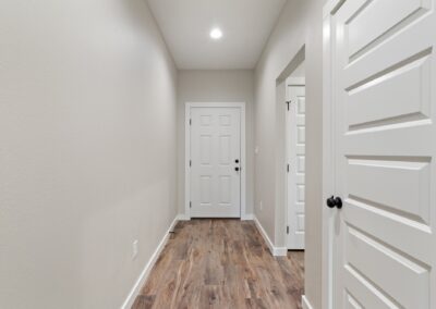 A hallway with light beige walls, wooden flooring, white doors, and recessed ceiling light. Two doors are visible on the right, and one is at the end of the hall.
