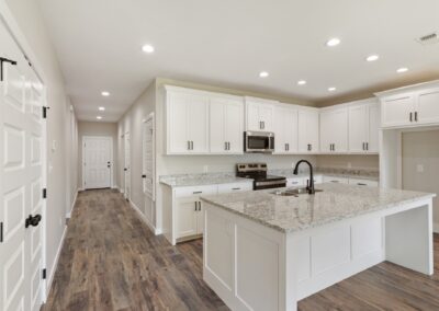 Modern kitchen with white cabinets, granite countertops, stainless steel appliances, an island with a sink, wood floors, and recessed ceiling lights. A hallway with doors is visible to the left.