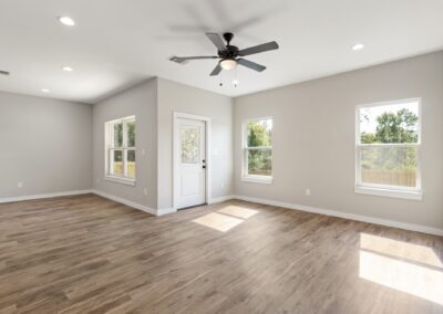 Bright, empty room with light wood flooring, beige walls, three windows, a white door, and a black ceiling fan. Sunlight streams through the windows, illuminating the spacious interior.
