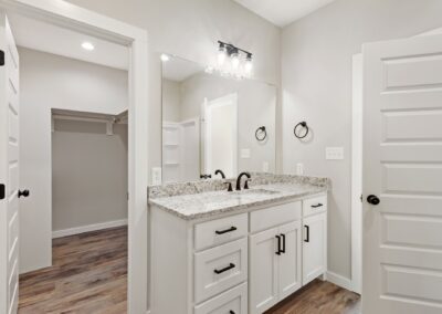 Modern bathroom with white cabinets, granite countertop, black fixtures, a large mirror, and wood flooring. An open door reveals a walk-in closet with shelves and a hanging rod. The space is brightly lit.