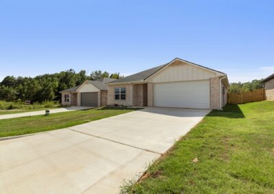 Single-story house with a brick and light siding exterior, two-car garage, and wide driveway. The home is set on a green lawn with a clear blue sky and neighboring houses nearby.