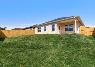 A single-story brick house with a sloped grassy backyard and wooden fence under a clear blue sky.