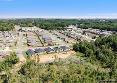 Aerial view of a suburban neighborhood with rows of houses, streets, and driveways bordered by green trees and a forested area under a clear blue sky.