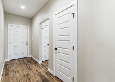 A hallway with light-colored walls, wood flooring, and three white doors with black handles. The area is well-lit with recessed lighting in the ceiling.
