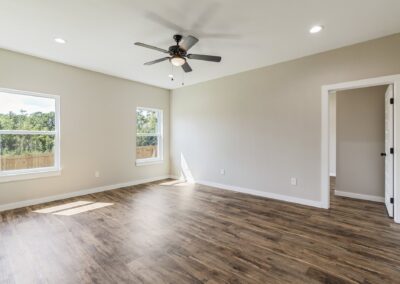 Empty room with light-colored walls, two windows, wood flooring, ceiling fan with light, recessed lighting, and an open door leading to another room. Sunlight streams in through the windows.