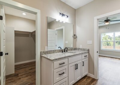 Modern bathroom with a granite countertop vanity, white cabinets, and a large mirror. Open doorways lead to a walk-in closet and a bedroom with wood floors and a ceiling fan.